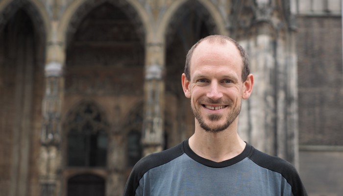 Smiling man standing outdoors with historic stone building background.